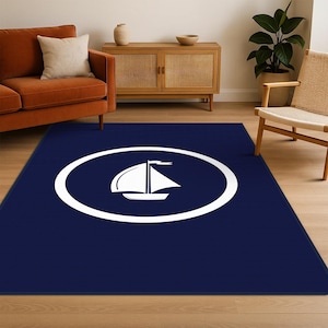 May include: A rectangular navy blue rug featuring a white sailboat design inside a white oval. The rug is placed on a hardwood floor in a living room setting, with a sofa, chair, and wooden cabinet in the background.