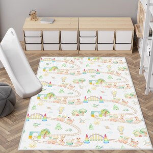 May include: A white play mat with a colorful train track design featuring trains, trees, and bridges. The mat is designed for children and is placed on a wooden floor. A white slide is visible on the left.