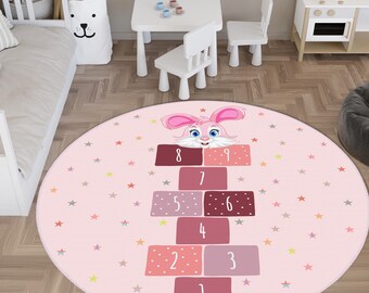 May include: A round pink children's rug with a hopscotch design. The rug features a cartoon bunny at the top and numbers 1-9 in a hopscotch layout. Colorful stars are scattered across the rug's surface.