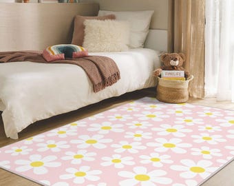May include: A rectangular pink rug featuring a pattern of white daisies with yellow centres. The rug is placed in a bedroom setting, with a bed, pillows, and a teddy bear in the background. The rug's dimensions are not visible.