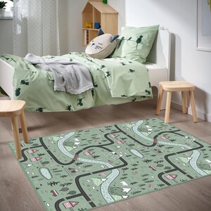 May include: A child's bedroom scene featuring a light green bed set with cat silhouettes and a matching road map rug. The rug has a road design with houses, trees, and mountains. Two small wooden stools and a wooden shelf are also visible.