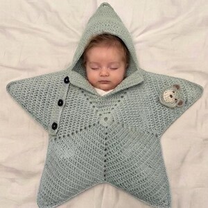 May include: A light sage green star-shaped crochet baby swaddle blanket with a hood. The garment features three black buttons along one side and a small, embroidered bear face appliqué on one of the star points, providing a cozy wrap for an infant.
