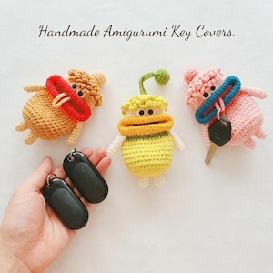 May include: Three handmade amigurumi key covers featuring crocheted characters with wide mouths. One is tan with a red mouth, one is yellow with a green sprout and yellow mouth, and one is pink with a blue mouth holding a metal key.