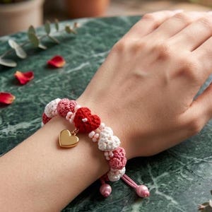Peut inclure: Un bracelet en crochet avec des perles en forme de cœur rouges, blanches et roses. Un charm en forme de cœur doré est suspendu au bracelet. Le bracelet est sur le poignet d'une personne.