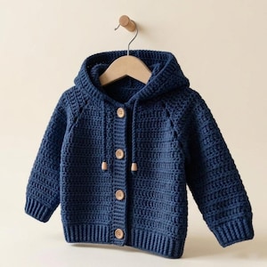 May include: A navy blue, hooded crochet cardigan with wooden buttons and drawstrings. The baby sweater is displayed on a wooden hanger against a neutral background. The crochet stitch creates a textured appearance.