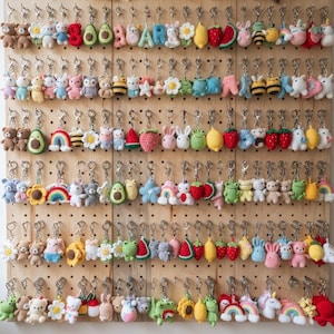 May include: A collection of colorful crochet keychains hanging on a wooden pegboard. The keychains feature various shapes including animals, fruits, flowers, rainbows, and letters, each attached to a silver metal ring and clasp.