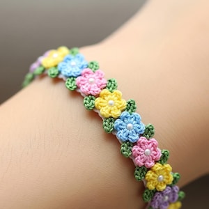 Op de afbeelding: Een gehaakte armband met een bloemenpatroon. De armband heeft kleine, kleurrijke bloemen in geel, blauw, roze en paars, met groene bladeren. Elke bloem heeft een kleine witte kraal in het midden. De armband heeft een witte basis.
