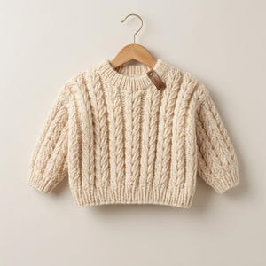 Puede incluir: Suéter de punto trenzado color crema con cuello redondo y mangas largas. El suéter se muestra en una percha de madera. Una pequeña etiqueta de cuero está adherida al suéter. La prenda de punto presenta un patrón de cable vertical texturizado.