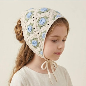 May include: A cream crochet bonnet with blue rose and green leaf detailing. The bonnet has a scalloped edge and ties under the chin. This handmade bonnet is designed for children. The bonnet is a traditional style.