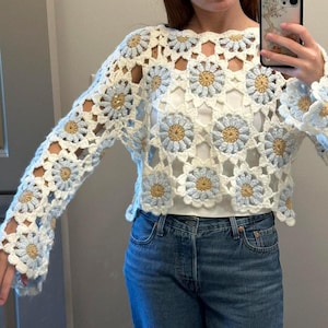 Könnte beinhalten: Ein weißes Häkeltop mit einem Gänseblümchenmuster in Hellblau und Gold. Das langärmlige Oberteil hat einen runden Ausschnitt und wird mit einer blauen Jeans kombiniert. Das Gänseblümchenmuster wiederholt sich im gesamten Design des Tops.