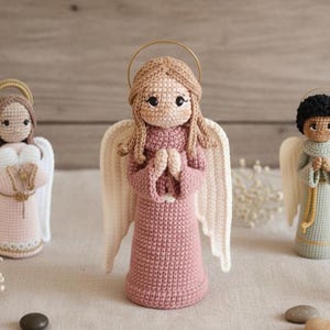 Puede incluir: Tres figuras de ángeles de ganchillo hechas a mano. Los ángeles tienen piel clara, cabello rubio y negro, y visten vestidos en rosa, blanco y verde. Cada ángel tiene alas blancas y un halo dorado. Los ángeles están sobre una superficie clara.