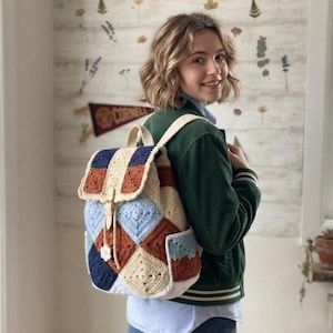 Puede incluir: Una mochila de ganchillo hecha a mano construida con motivos cuadrados en color crema, azul marino, azul claro y naranja óxido. La bolsa cuenta con una solapa superior con cierre de correa de cuero color canela claro, cordón y dos bolsillos laterales.