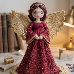 Puede incluir: Una muñeca de ángel hecha a mano con ganchillo, con un vestido rojo y alas doradas. La muñeca tiene cabello castaño y un rostro delicado. El vestido presenta mangas acampanadas y un dobladillo festoneado. Las alas están intrincadamente tejidas. Un artículo decorativo.