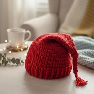May include: A handmade, red crochet hat with a long, pointed end and a tassel. The hat is sitting on a white surface, with a cup of tea and decorative lights in the background. The hat is a winter accessory.