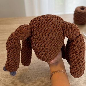 Puede incluir: Un pavo de ganchillo marrón hecho a mano con las alas extendidas y pequeños detalles azules en los pies. El pavo de peluche está hecho con un hilo grueso. Se ve un ovillo de hilo marrón en el fondo.