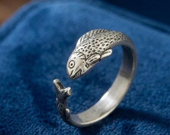 Op de afbeelding: Een zilverkleurige ring met een visontwerp. De vis is gedetailleerd met schubben en een oog, waarbij het uiteinde van de staart de opening van de band vormt. De ring staat tegen een blauwe achtergrond.