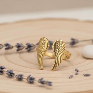 Puede incluir: Un anillo dorado con un dise&ntilde;o de dos alas estilizadas. El anillo est&aacute; sobre una superficie de madera clara, con ramitas de lavanda y una peque&ntilde;a piedra blanca en el fondo. Las alas tienen patrones de plumas detallados.