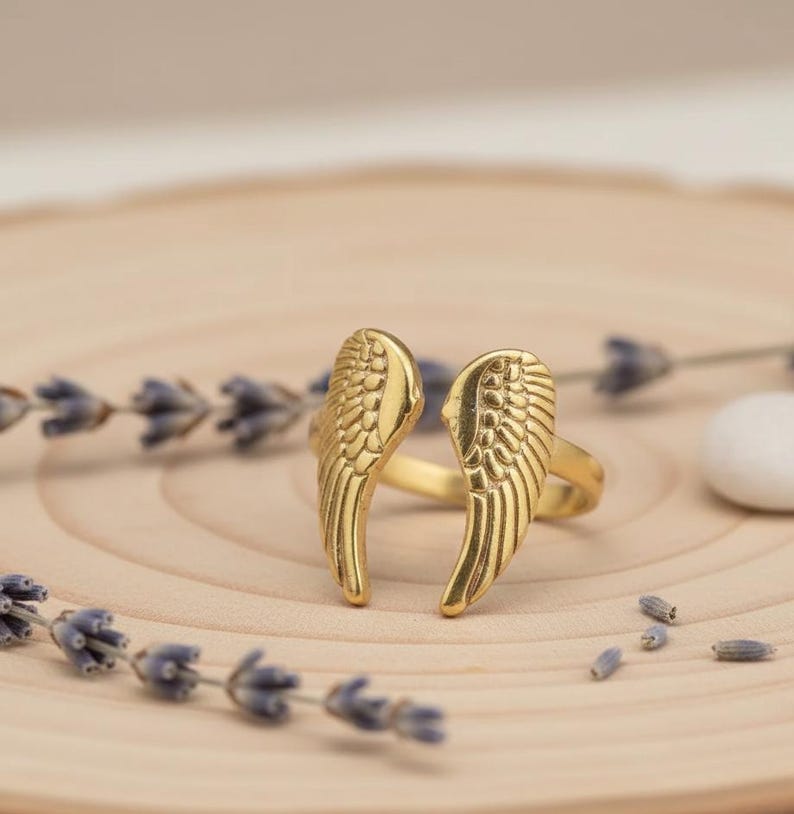 Puede incluir: Un anillo dorado con un dise&ntilde;o de dos alas estilizadas. El anillo est&aacute; sobre una superficie de madera clara, con ramitas de lavanda y una peque&ntilde;a piedra blanca en el fondo. Las alas tienen patrones de plumas detallados.