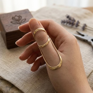 May include: A gold ring with a distinctive design, worn on a finger. The ring features a band at the top of the finger and a long, curved element running down the side. A wooden box with a floral motif is in the background.