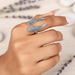 Puede incluir: Un anillo plateado con dise&ntilde;o de alas. El anillo envuelve el dedo con dos formas de alas detalladas, una arriba y otra abajo. El anillo se muestra en una mano con un fondo claro.