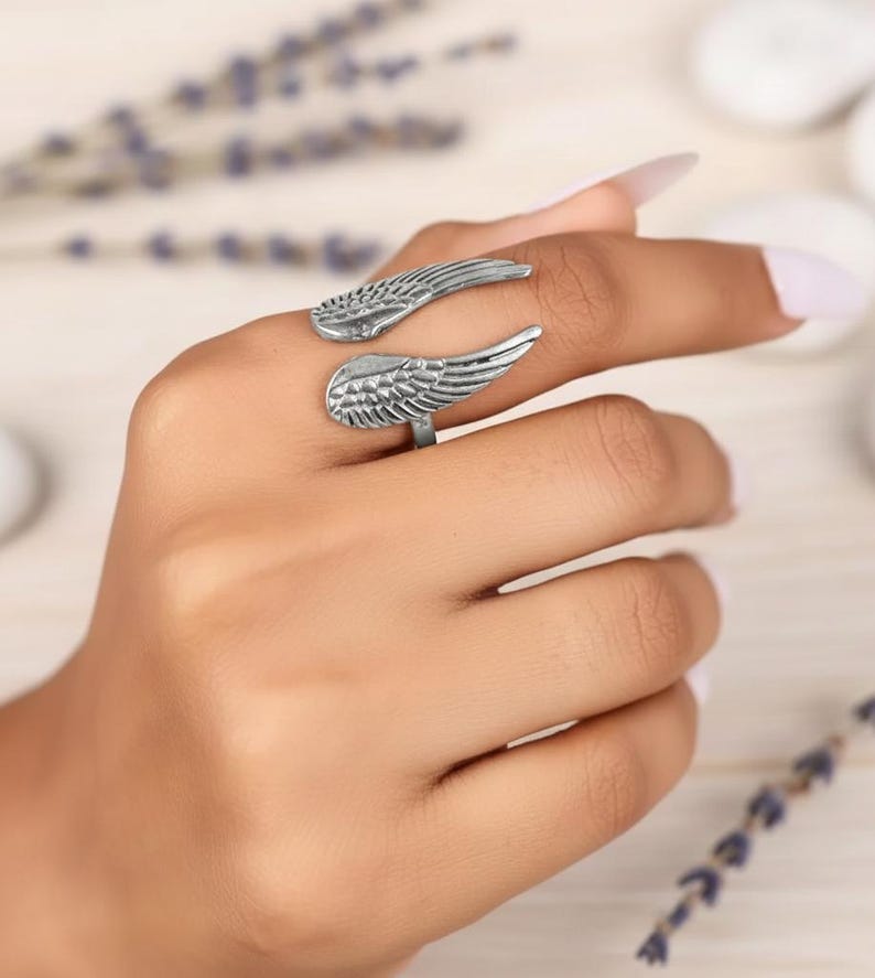 Puede incluir: Un anillo plateado con dise&ntilde;o de alas. El anillo envuelve el dedo con dos formas de alas detalladas, una arriba y otra abajo. El anillo se muestra en una mano con un fondo claro.
