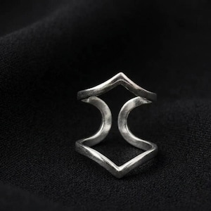 May include: A silver-toned ring with a geometric design. The ring features two chevron shapes connected by a curved bar. The ring is set against a black background.