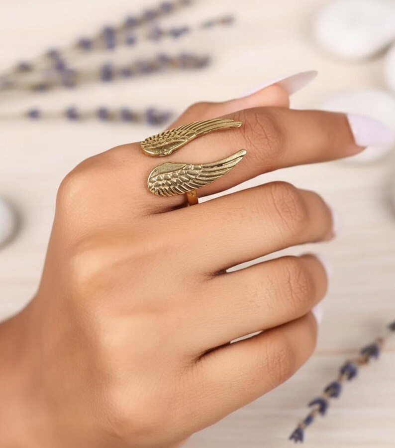 Puede incluir: Un anillo dorado con un dise&ntilde;o de alas. El anillo se envuelve alrededor del dedo con dos formas de alas detalladas. El anillo se muestra en una mano con un fondo de color claro.