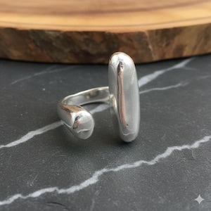 Peut inclure: Une bague en argent au design unique. La bague présente un anneau arrondi qui épouse le doigt, avec un grand élément vertical argenté et allongé. La bague est posée sur une surface sombre et texturée.