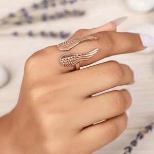 Puede incluir: Un anillo de oro rosa con dise&ntilde;o de alas. El anillo se envuelve alrededor del dedo con texturas detalladas de plumas en cada ala. La joya se muestra en una mano, mostrando el dise&ntilde;o y el color del anillo.