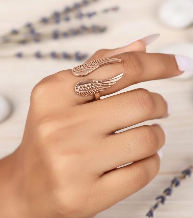 Puede incluir: Un anillo de oro rosa con dise&ntilde;o de alas. El anillo se envuelve alrededor del dedo con texturas detalladas de plumas en cada ala. La joya se muestra en una mano, mostrando el dise&ntilde;o y el color del anillo.
