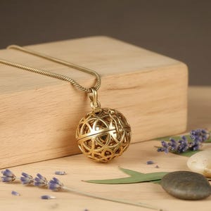 Puede incluir: Un collar con colgante dorado con un diseño esférico calado. El intrincado patrón del colgante cuelga de una delicada cadena. El collar descansa sobre una superficie de madera, con ramitas de lavanda y pequeñas piedras cerca.