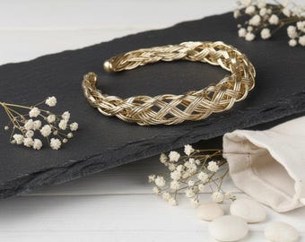 Puede incluir: Brazalete dorado con diseño trenzado. El brazalete abierto se exhibe sobre una superficie de pizarra negra, acentuado con flores blancas y pequeñas piedras blancas. Se destaca el intrincado patrón del brazalete.