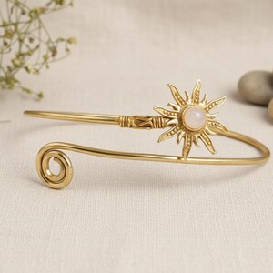 May include: A gold arm cuff with a sunburst design and a central, pale stone. The cuff has a spiral detail at one end and a decorative element near the sunburst. The jewellery is displayed on a neutral-toned surface.