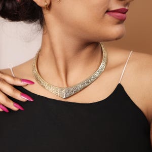 May include: A silver-toned collar necklace with an intricate floral pattern. The necklace has a V-shaped design that sits around the neck. The jewelry is worn over a black top.