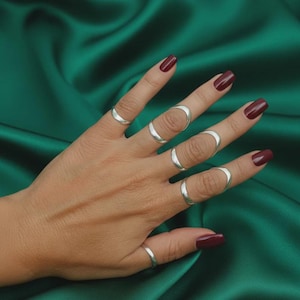 Peut inclure: Une main ornée de plusieurs bagues en argent de différents modèles, dont une bague qui recouvre tout le doigt. Les ongles sont peints en rouge foncé. Le fond est un tissu vert foncé.
