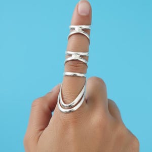 May include: A hand wearing multiple silver rings against a blue background. The rings include a double-banded ring, a simple band, and several rings with a bridge design. The rings are made of shiny metal.