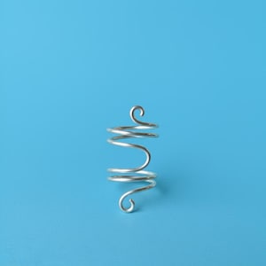 May include: A silver-colored, spiral-shaped ring with a unique design. The ring features a series of curved lines and loops, creating an abstract and artistic appearance. The ring is set against a solid blue background.