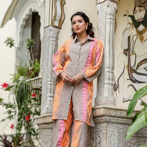 Könnte beinhalten: Ein zweiteiliges Leinen-Outfit in neutralem Grau, akzentuiert mit leuchtenden orangefarbenen und rosa geometrischen Mustern. Das Ensemble umfasst ein Hemd mit Knöpfen und eine passende Hose mit weitem Bein. Das Outfit wird von einer Person vor einer verzierten Wand getragen.