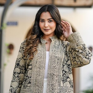 May include: A black and beige patterned jacket with a mandarin collar. The jacket features an intricate floral design and is worn over a white top. The sleeves are long, and the jacket is open in the front.