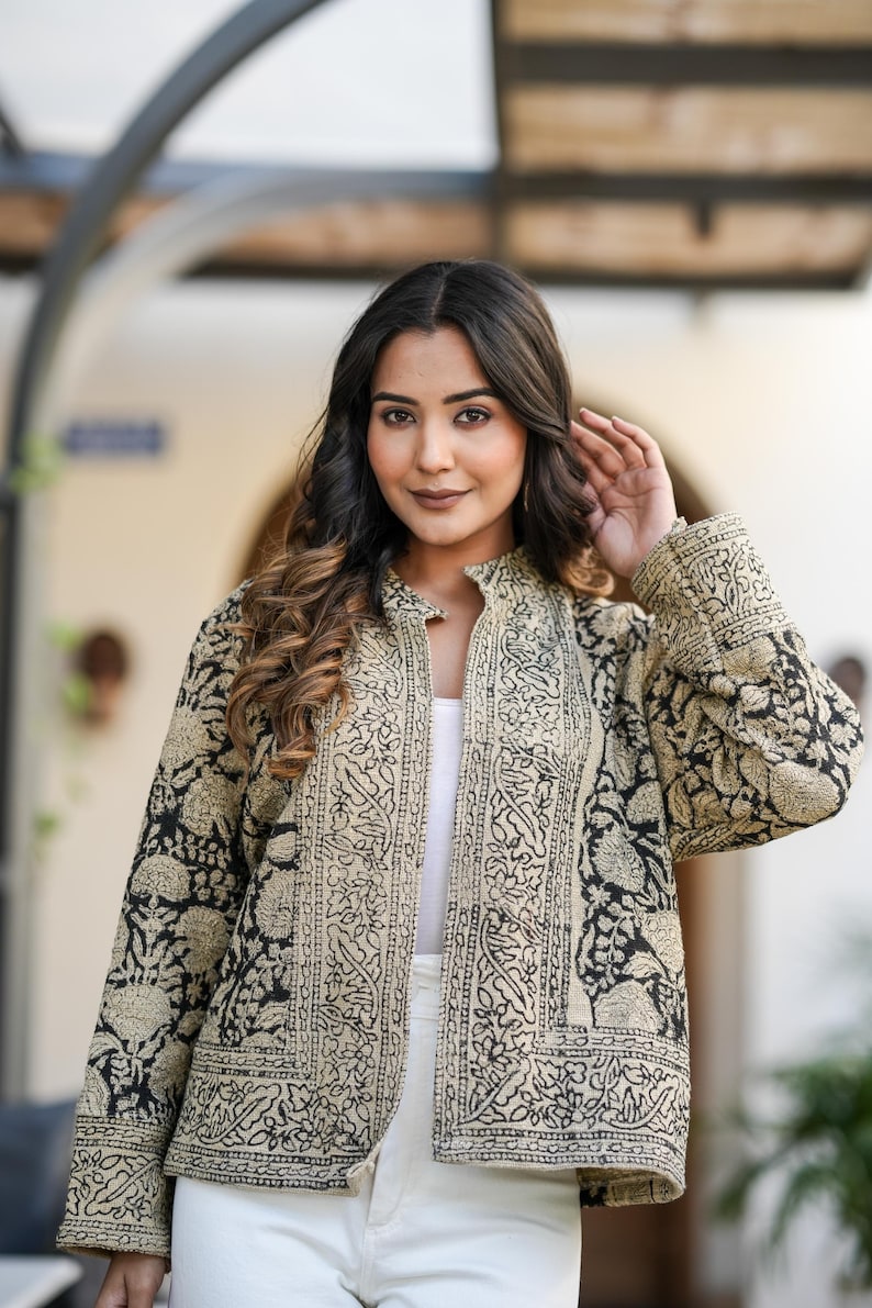 May include: A black and beige patterned jacket with a mandarin collar. The jacket features an intricate floral design and is worn over a white top. The sleeves are long, and the jacket is open in the front.