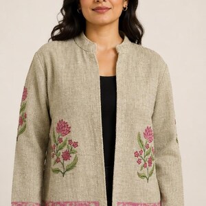 Könnte beinhalten: Eine beigefarbene, vorne offene Jacke mit Stehkragen und langen Ärmeln. Das Kleidungsstück ist mit gestickten rosa Blumenmotiven auf den Vorderteilen und Ärmeln verziert, ergänzt durch eine gemusterte rosa Borte am unteren Saum.