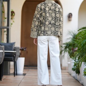 May include: A black and beige floral print jacket with long sleeves and white trousers. The jacket features a detailed pattern and is paired with wide-leg white trousers. The outfit is completed with heeled shoes.