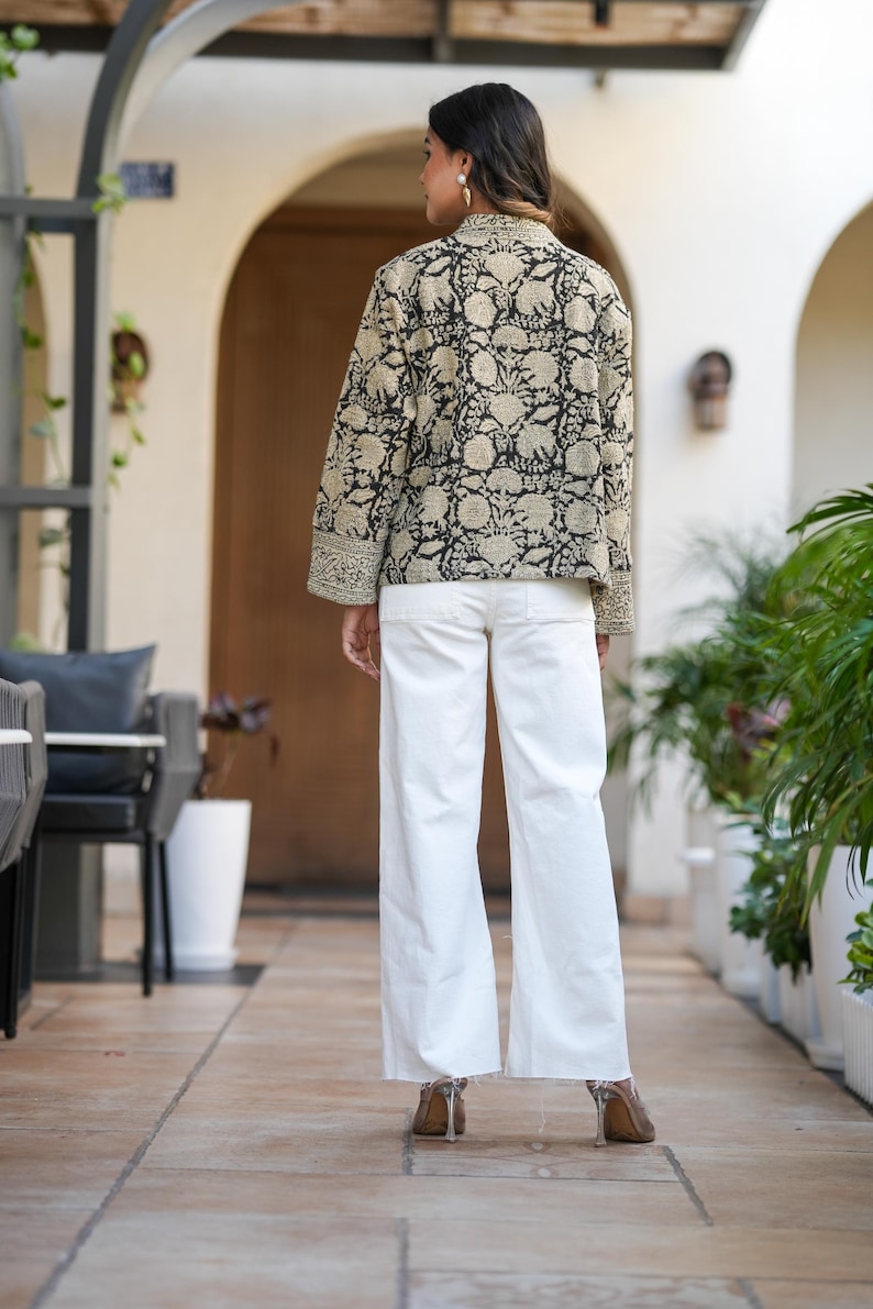 May include: A black and beige floral print jacket with long sleeves and white trousers. The jacket features a detailed pattern and is paired with wide-leg white trousers. The outfit is completed with heeled shoes.