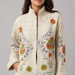 Può includere: Giacca color crema con ricami floreali arancioni, verdi e viola. La giacca ha un collo alto, maniche lunghe e un tessuto strutturato. Il design floreale è sul davanti e sulle maniche.