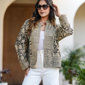 May include: A beige and black patterned jacket with a mandarin collar. The jacket is worn over a white top and paired with white trousers. The jacket's design features intricate floral and geometric patterns. The model is wearing sunglasses.