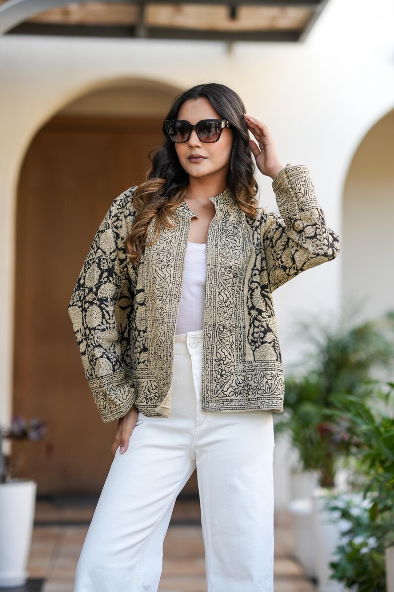 May include: A beige and black patterned jacket with a mandarin collar. The jacket is worn over a white top and paired with white trousers. The jacket's design features intricate floral and geometric patterns. The model is wearing sunglasses.
