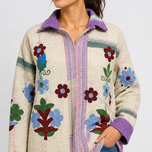 Könnte beinhalten: Cremefarbene Jacke mit floralen Stickereien in Rot, Blau und Grün. Die Jacke hat einen lila Kragen und Manschetten mit vertikalen Streifen in Rosa und Grün. Eine stilvolle, bestickte Jacke für den Alltag.