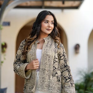 May include: A beige and black patterned jacket with a mandarin collar. The jacket features an intricate floral design and is worn over a white top and white trousers. The jacket is a lightweight, stylish garment.