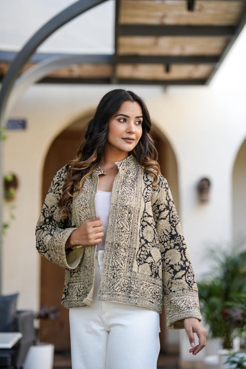 May include: A beige and black patterned jacket with a mandarin collar. The jacket features an intricate floral design and is worn over a white top and white trousers. The jacket is a lightweight, stylish garment.