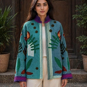 Könnte beinhalten: Türkisfarbene Jacke mit Blumen- und Blättermuster, lila Kragen und Manschetten. Die Jacke wird über einem weißen Oberteil und cremefarbenen Hosen getragen. Der Hintergrund zeigt Pflanzen in Terrakotta-Töpfen.